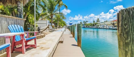 A wooden dock leading to a calm body of water, with a thatched-roof structure and palm trees on the left.