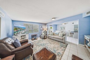 Ein Wohnzimmer mit Sofa, Sessel, Hocker, Fernseher, einem Gemälde und Blick ins Freie.