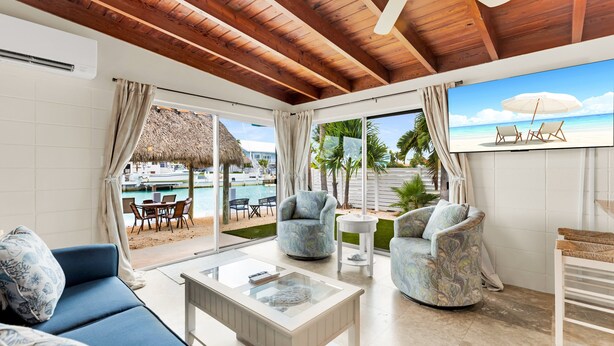 A spacious living area with a wooden ceiling, a large TV screen, a blue sofa, two patterned armchairs, and a glass coffee table. - Reel-Axed Retreat ~ Pet Friendly, 37.5' Dock, Tiki Hut, Cabana Club w Pool Access (Key Colony Beach)