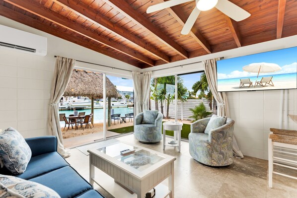 A spacious living area with a wooden ceiling, a large TV screen, a blue sofa, two patterned armchairs, and a glass coffee table. - Reel-Axed Retreat ~ Pet Friendly, 37.5' Dock, Tiki Hut, Cabana Club w Pool Access (Key Colony Beach)