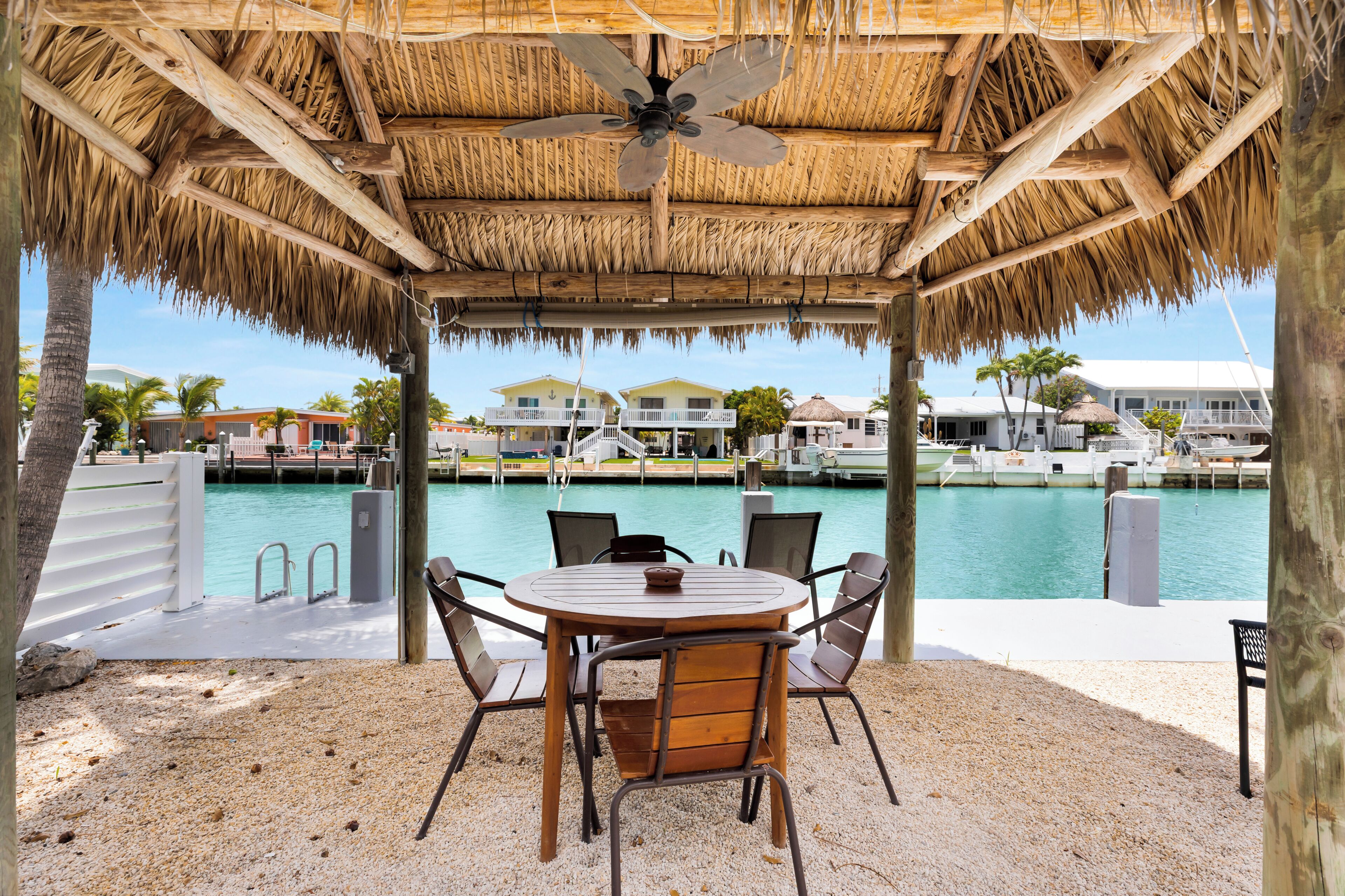 A covered outdoor seating area with a table and chairs, a thatched roof, and a view of a marina with boats and houses.