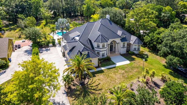 A large, two-story house with a dark roof, surrounded by lush greenery and a swimming pool.