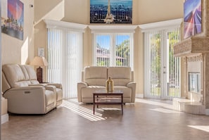 A spacious living room with a reclining chair, a sofa, a glass coffee table, and large windows with blinds.