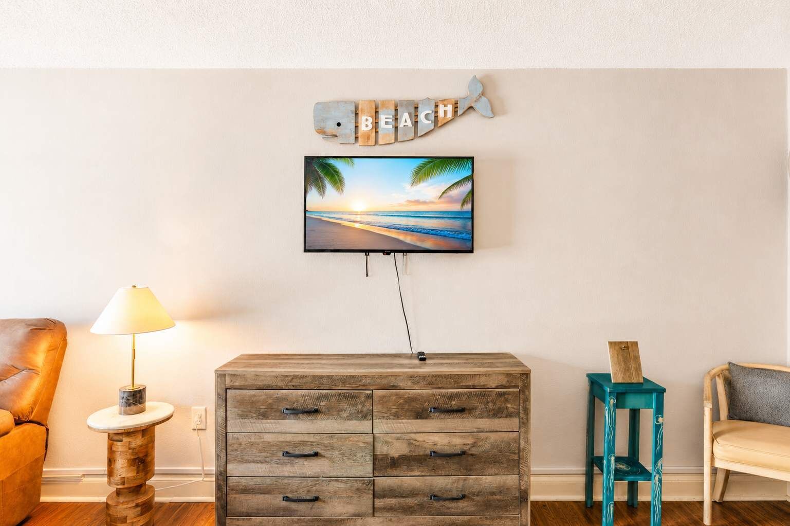 Een woonkamer met een aan de muur bevestigde televisie, een houten kast met lades, een lamp en twee stoelen.