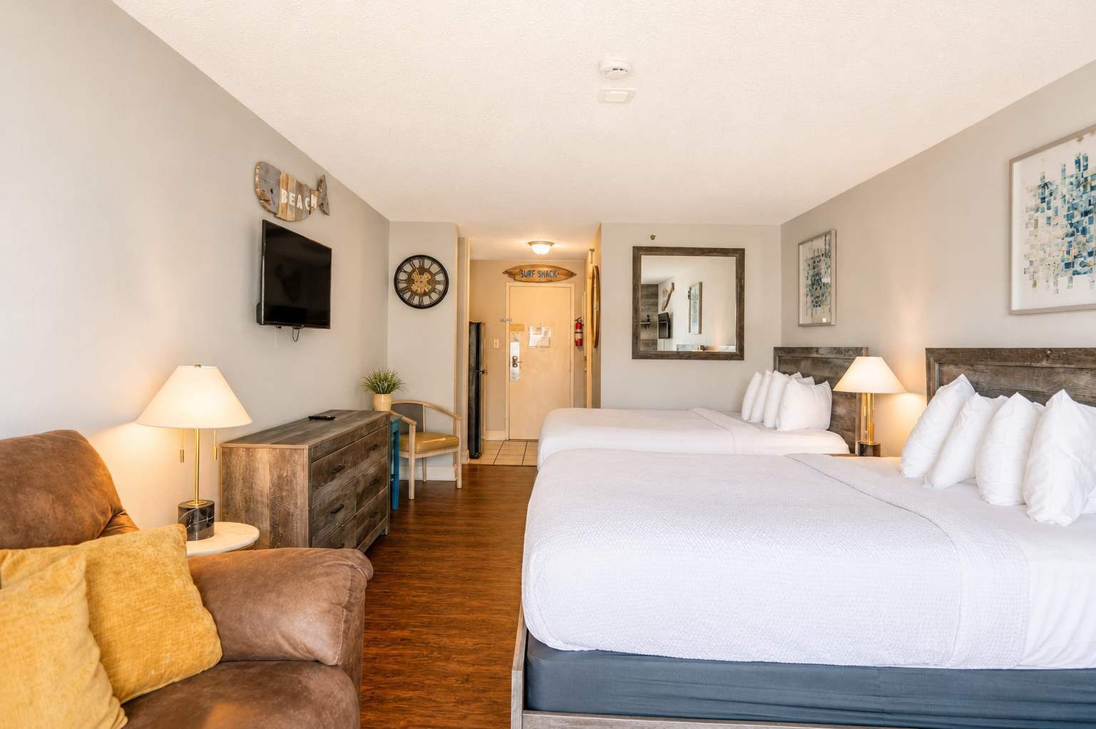 Une chambre d’hôtel comprenant un lit, deux tables de chevet, un téléviseur, une horloge, un miroir et une porte donnant accès à une salle de bain.