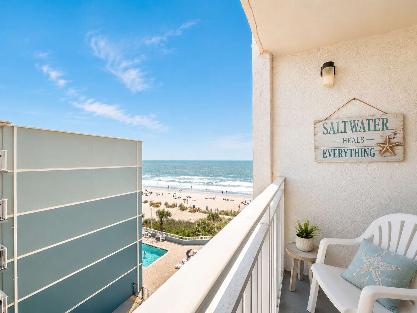 Ein Balkon mit Blick auf den Strand, ein Schild mit der Aufschrift 'Saltwater heals everything', ein kleiner Tisch mit einer Pflanze und ein weißer Stuhl mit Kissen in Form eines Seesterns.
