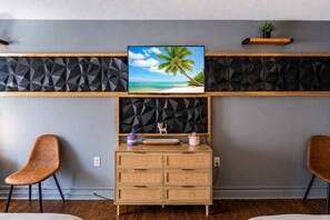 Ein modernes Wohnzimmer mit einem an der Wand mit geometrischem Muster befestigten Fernseher, einer Holzkassette mit Schubladen, zwei braunen Sesseln und einem kleinen Regal mit einer Pflanze.