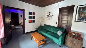 Une pièce avec un canapé vert, une table basse en bois, des œuvres d’art décoratives accrochées au mur, une lampe et une radio ancienne.