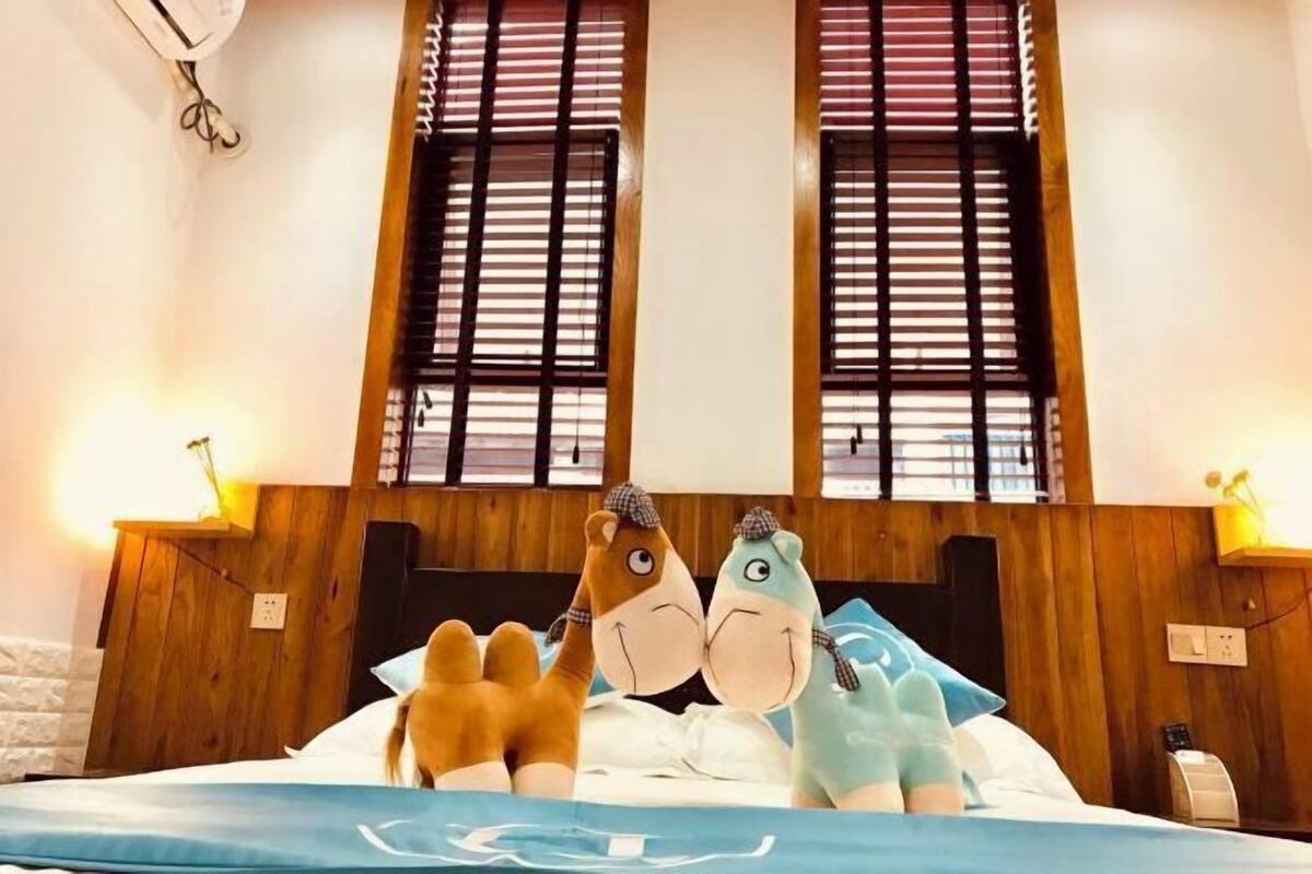 A bed with plush toys, wooden headboard, and windows with blinds.