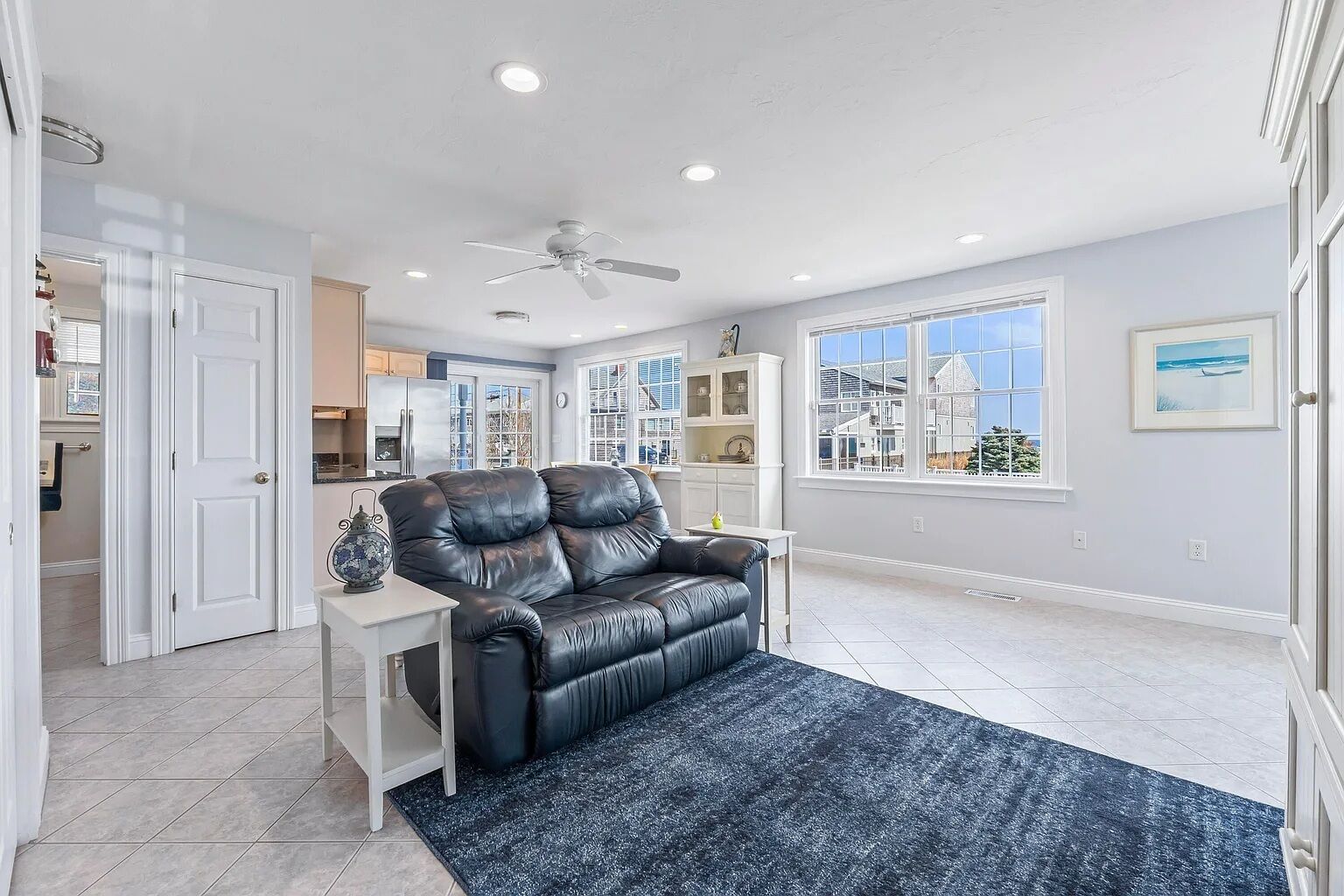 A living room with a black leather sofa, a white side table, a ceiling fan, and a view of the outside through large windows.