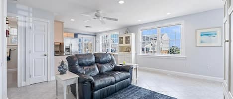 A living room with a black leather sofa, a white side table, a ceiling fan, and a view of the outside through large windows.