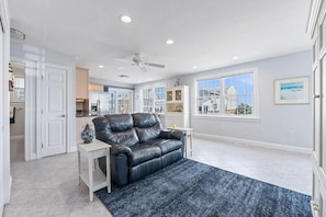 A living room with a black leather sofa, a white side table, a ceiling fan, and a view of the outside through large windows. - Amazing Beach House with Ocean Views - Just steps to Wells Beach (Wells)