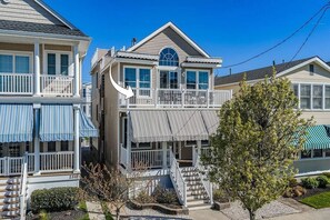 Une maison à deux étages avec un balcon, un porche à l’avant avec un escalier et un arbre dans la cour.