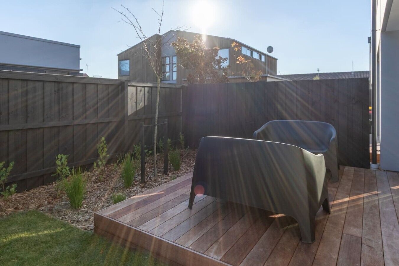 Eine Holzterrasse mit zwei Stühlen, ein eingezäunter Garten und ein Gebäude im Hintergrund.