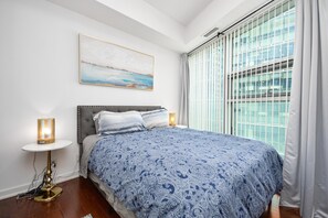 A bedroom with a large bed, a bedside table with a lamp, a window with blinds, and a painting on the wall. - Urban 1BR Escape with Queen Bed in Prime Toronto Location (Toronto)