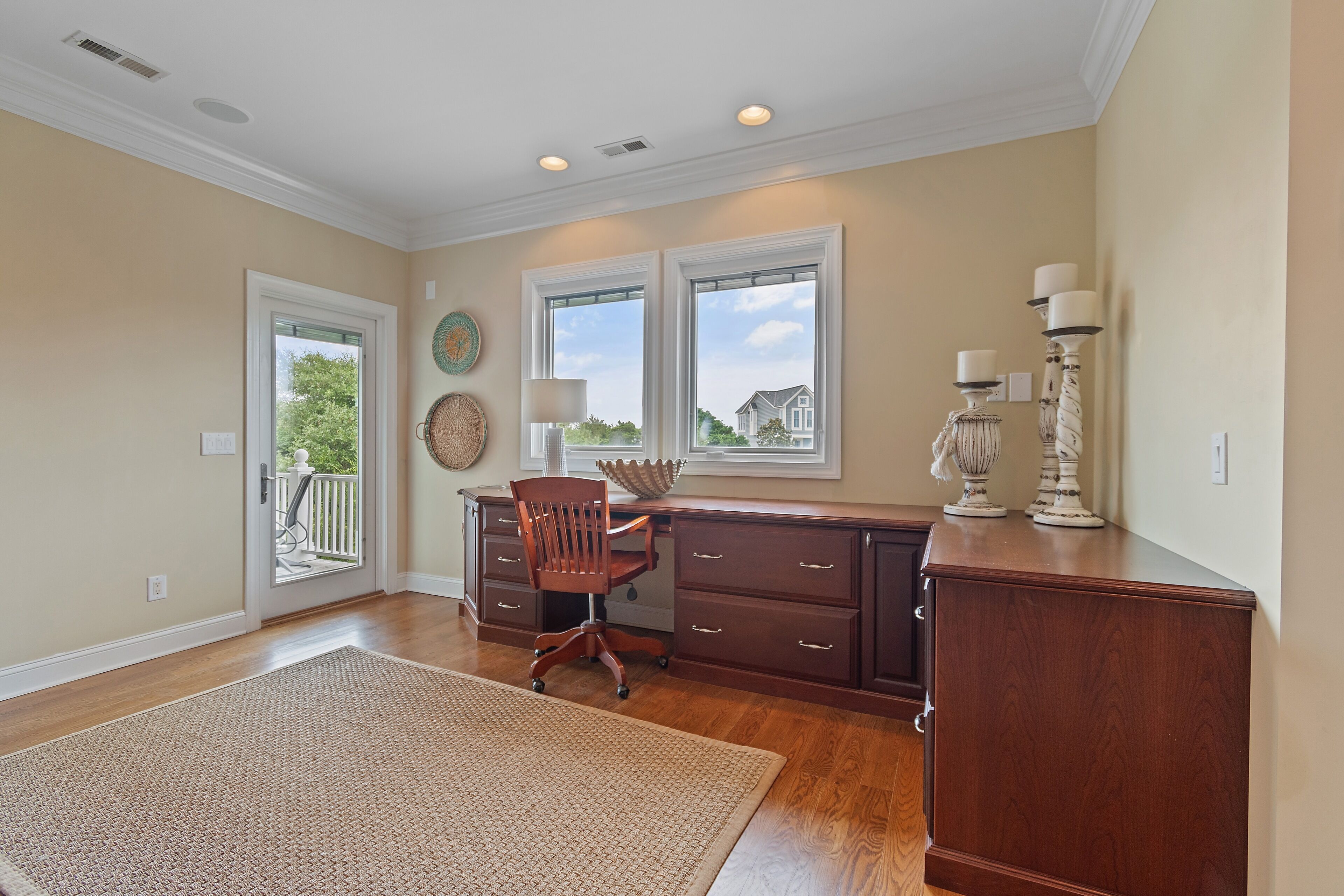 A room with a wooden desk, a chair, a large rug, a window with a view, and a door leading outside.