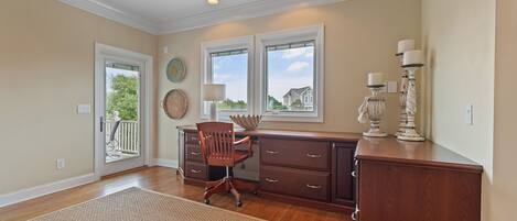 A room with a wooden desk, a chair, a large rug, a window with a view, and a door leading outside.