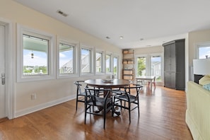 A spacious room with a wooden floor, a round dining table with chairs, a kitchen area with a refrigerator, and large windows offering views of greenery. - "Escape" 5 Bedrooms, 6.5 Bathrooms, ICW Home, Community Pool and Tennis Court! (Supply)