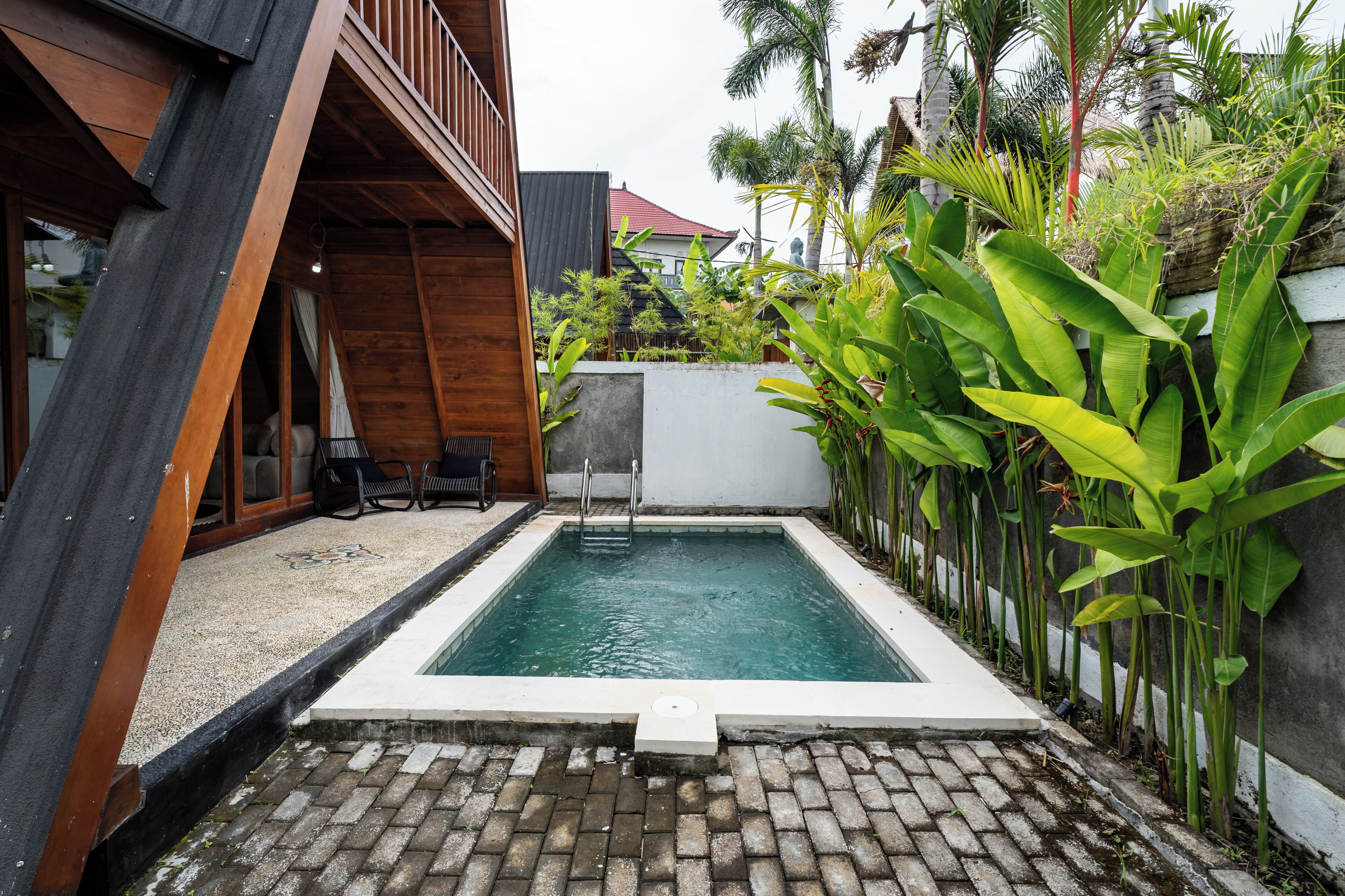 Una piscina rectangular con borde de hormigón, rodeada por un patio de ladrillo y una terraza de madera.