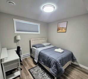 Ein Schlafzimmer mit Bett, Schreibtisch, Stuhl, Lampe, einem Fenster mit Jalousien, einem Gemälde und einem Heizkörper.