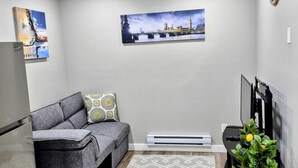 A living room with a gray sofa, a patterned throw pillow, a wall-mounted TV, and a framed picture of a bridge. - Femkem Cozy Suite in a peaceful and beautiful neighborhood. You will love it. (Regina)