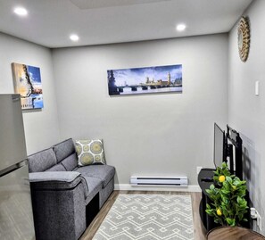 A living room with a gray sofa, a patterned throw pillow, a wall-mounted TV, and a framed picture of a bridge.