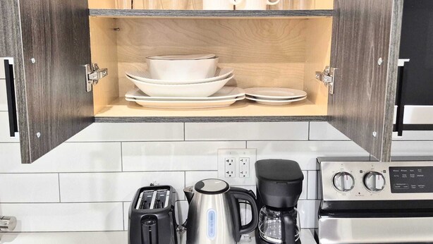 A kitchen with a wooden cabinet containing wine glasses, bowls, and plates. Below the cabinet, there is a toaster, a coffee maker, and a stove. - Femkem Cozy Suite in a peaceful and beautiful neighborhood. You will love it. (Regina)