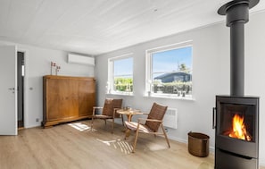Ein Zimmer mit einem Holzschrank, zwei Korbstühlen, einem kleinen Tisch, einem Kamin und großen Fenstern.