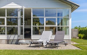 Ein modernes Haus mit großen Fenstern, einer Terrasse mit zwei Lounge-Stühlen und einem sichtbaren Innenbereich mit Essbereich.