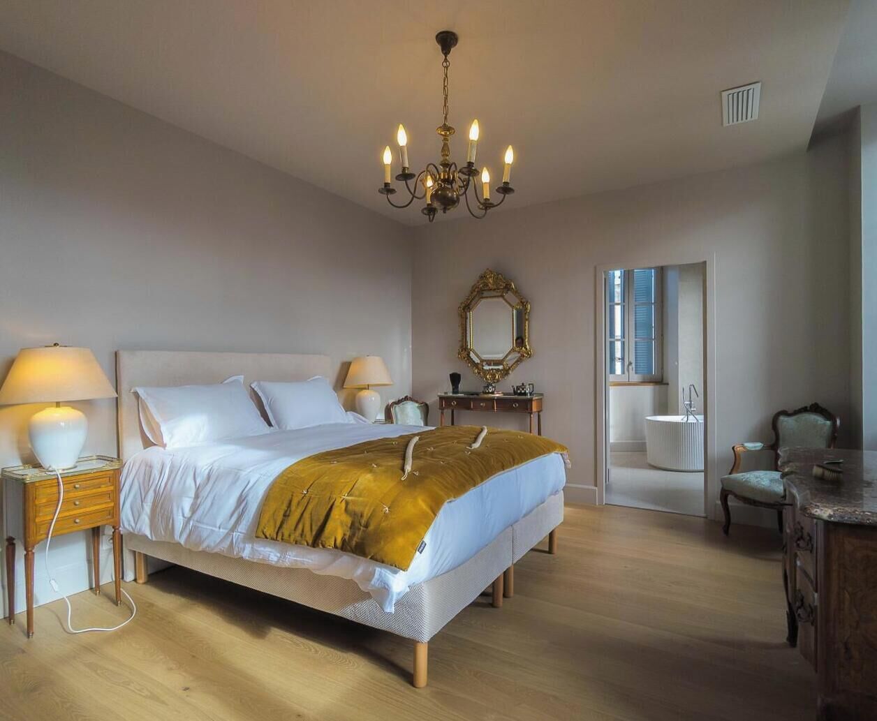 Une chambre à coucher comprenant un lit, des lampes de chevet, un lustre, un miroir, une chaise et une commode.