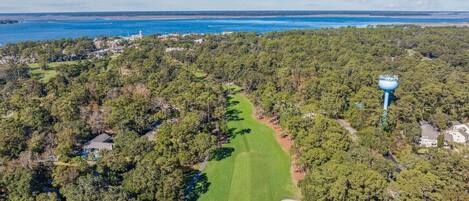 An aerial view of a golf course with a lighthouse, dense forest, and a coastal backdrop.