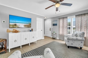 Ein Zimmer mit Deckenventilator, einem großen Fernseher, einem weißen Schrank, einem gemusterten Sessel und Meerblick.