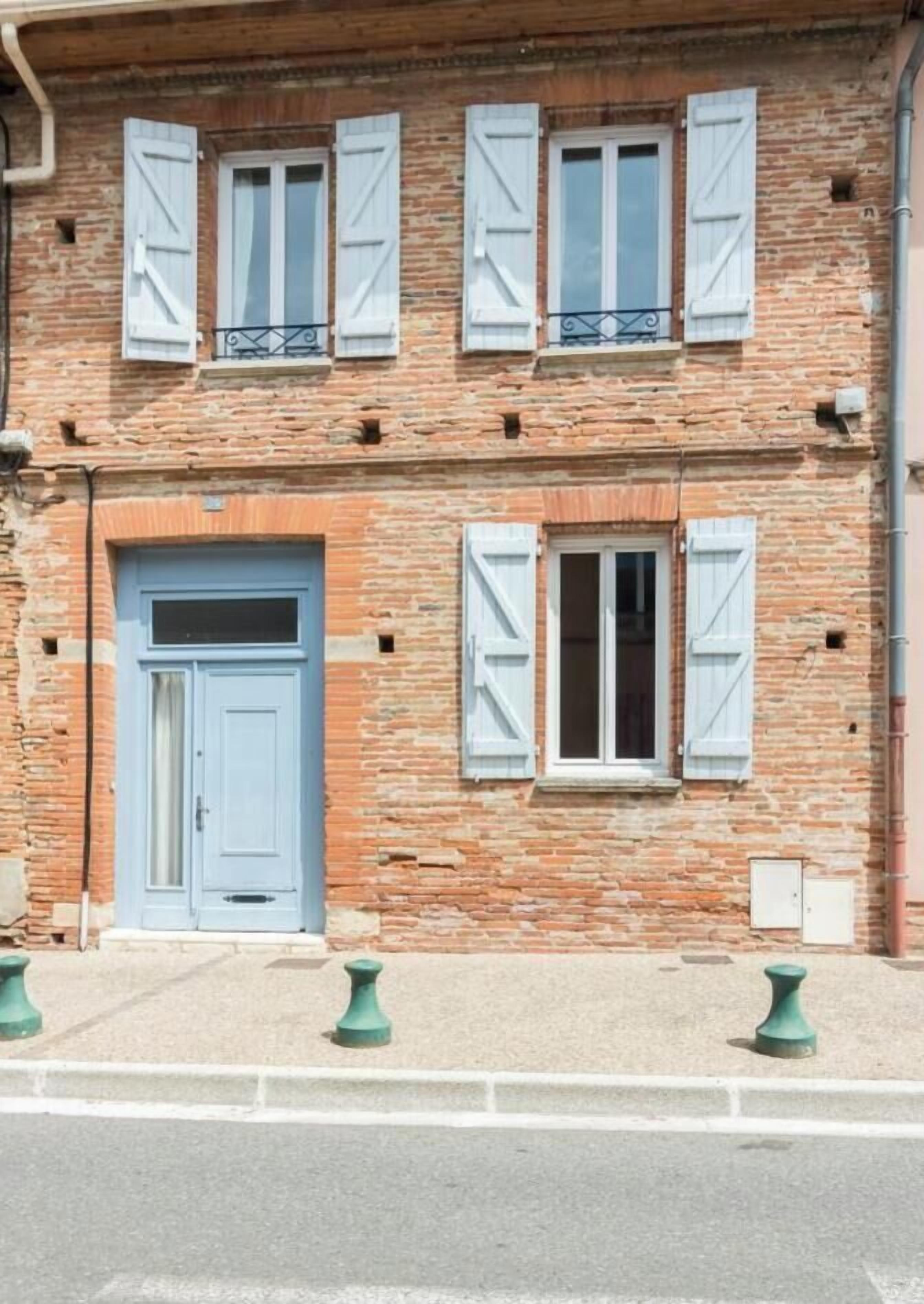 Un edifici de dues plantes amb una porta i persianes blaves, parets de maó i un petit balcó amb barana metàl·lica.