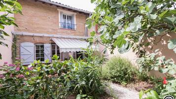 Une maison de campagne en briques, dotée d’un balcon, d’un porche abrité et d’un jardin où poussent diverses plantes et fleurs.
