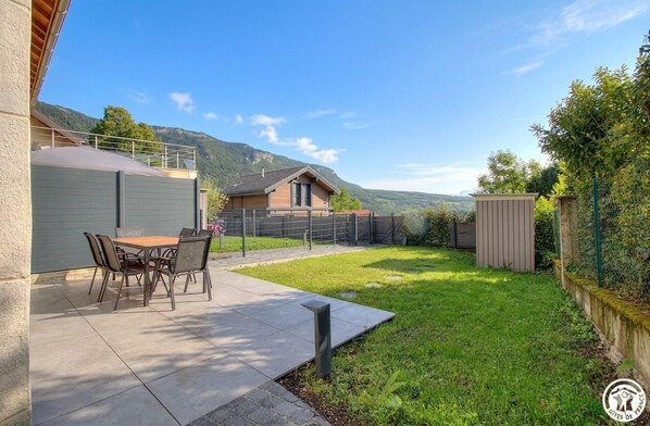 A patio with a table and chairs, a small house, and a fenced yard with a mountain backdrop.