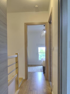 A hallway with a wooden floor, a door leading to a room with a window, and a ceiling light fixture.