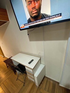 A small, modern room with a white desk, a chair, and a television mounted on the wall displaying a news broadcast. - Studio Copa Beach (Rio de Janeiro)