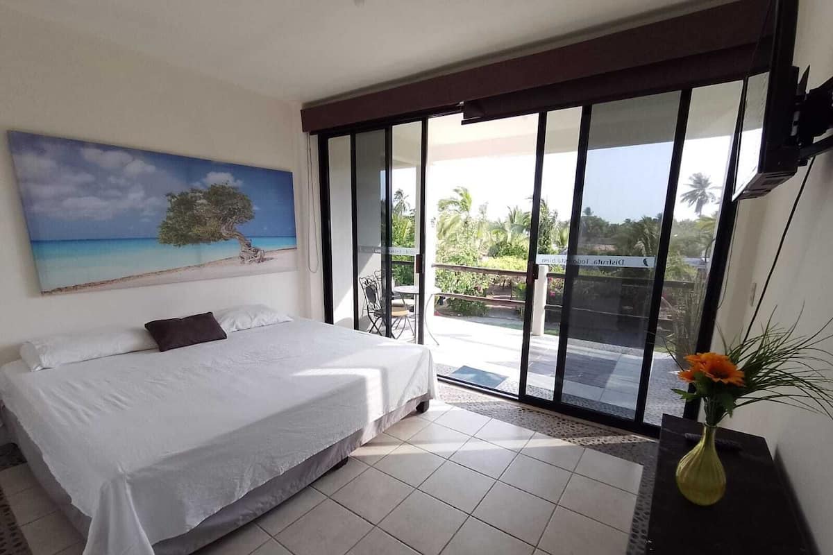 A bedroom with a bed, a painting of a beach, a sliding glass door to a balcony, and a vase with flowers.