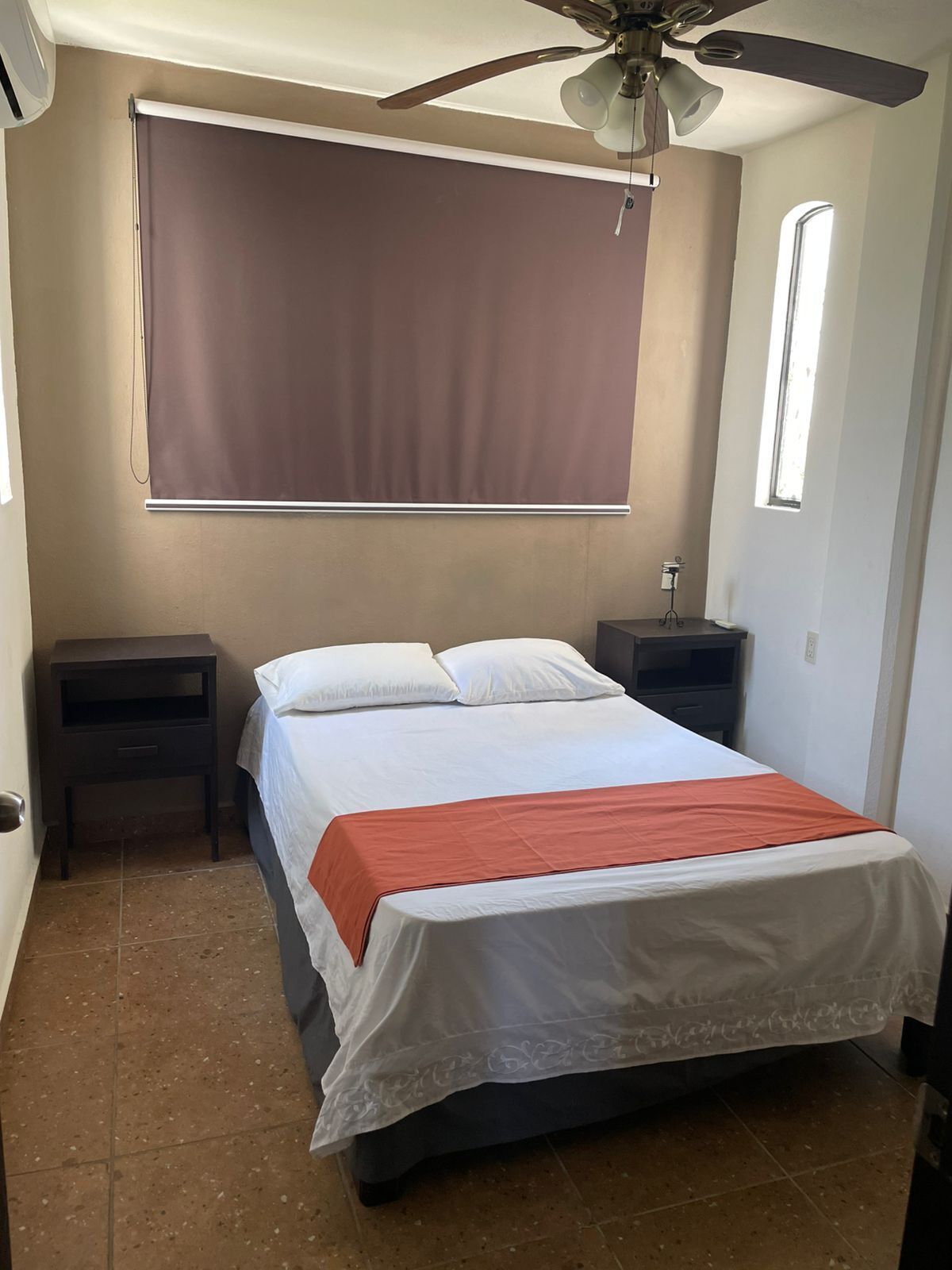 A bedroom with a bed, nightstands, a ceiling fan, a window, and a wall-mounted blind.