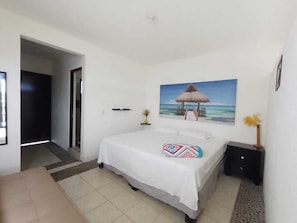 Un dormitorio con una cama, un cuadro de una playa, una mesita de noche y una puerta que da a otra habitación.