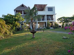 A two-story building with a thatched roof and a modern upper section, surrounded by lush greenery and a well-maintained lawn. - Zana Town Bungalows (Barra de Potosi)