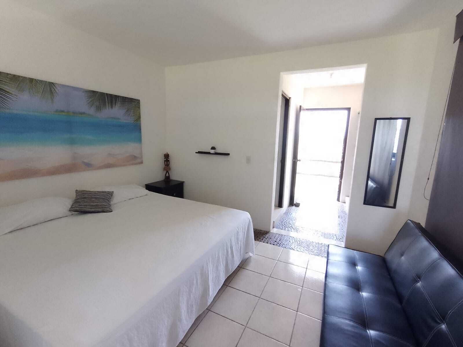 A bedroom with a bed, a bench, a painting, a nightstand, and a door leading to another room.