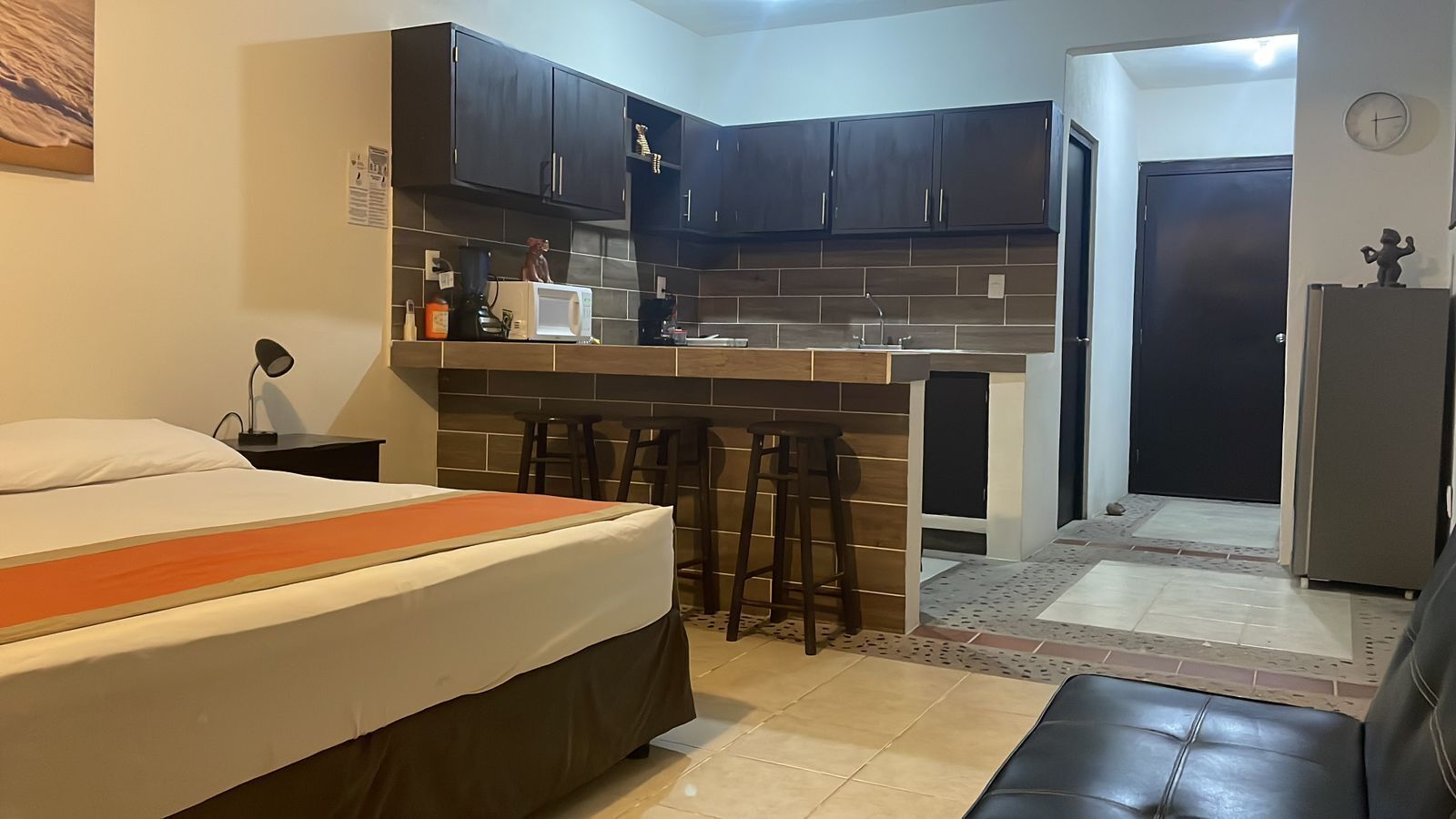 A compact hotel room with a bed, a kitchenette, a small bar area, a refrigerator, and a door leading to another room.