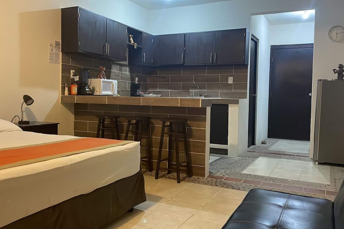 A compact hotel room with a bed, a kitchenette, a small bar area, a refrigerator, and a door leading to another room.