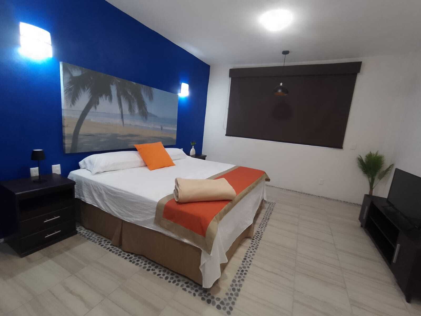 A bedroom with a bed, a nightstand, a lamp, a painting, a window, and a plant.