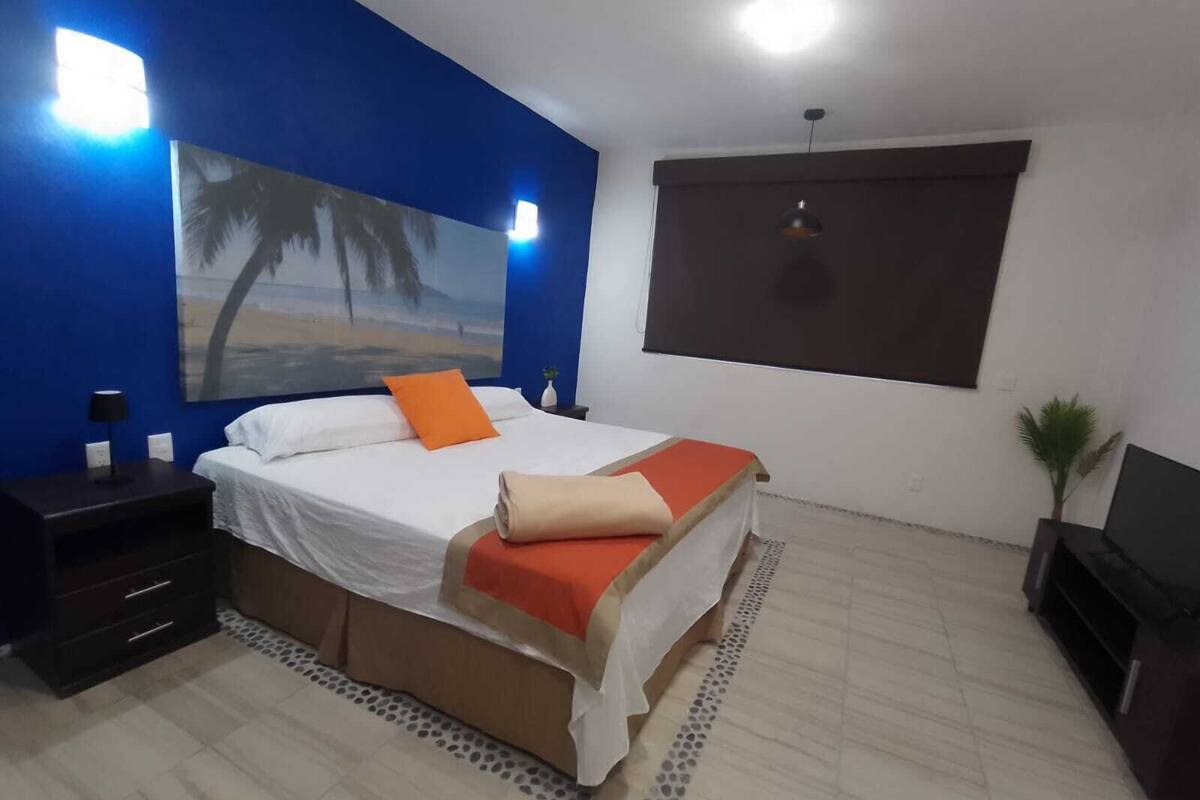 A bedroom with a bed, a nightstand, a lamp, a painting, a window, and a plant.
