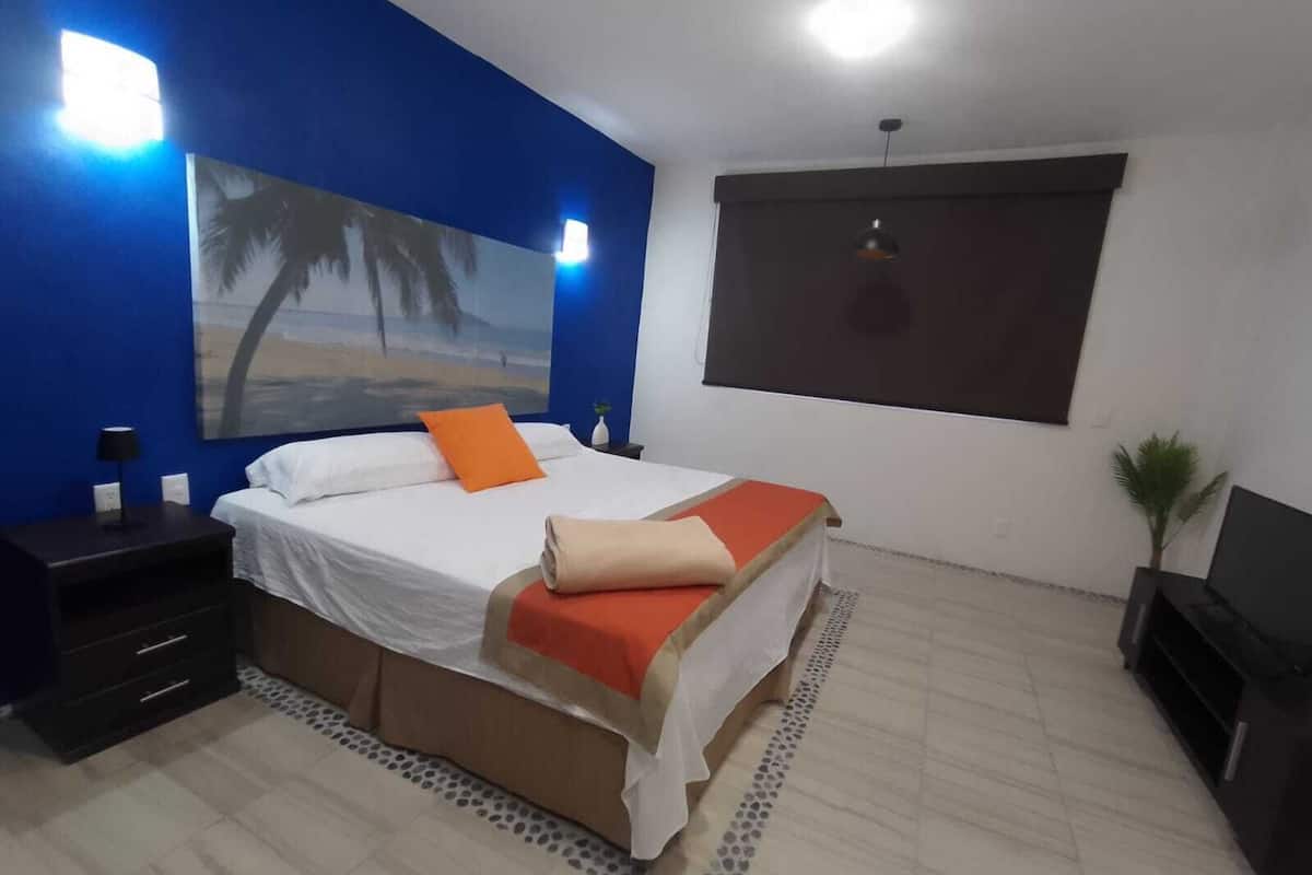 A bedroom with a bed, a nightstand, a lamp, a painting, a window, and a plant.