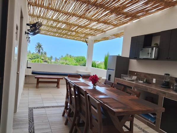 An outdoor kitchen with a wooden table and chairs, a built-in microwave, and a view of the greenery.
