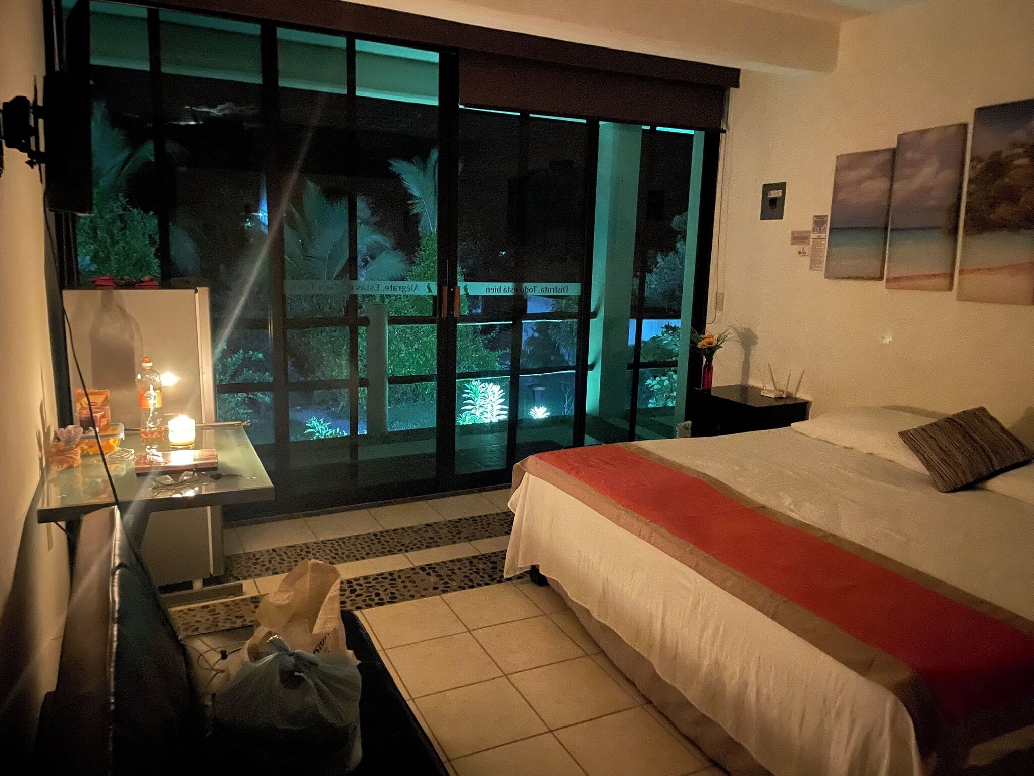 A hotel room with a bed, a desk, a chair, a table with a lamp, a vase with flowers, and a view through large windows.