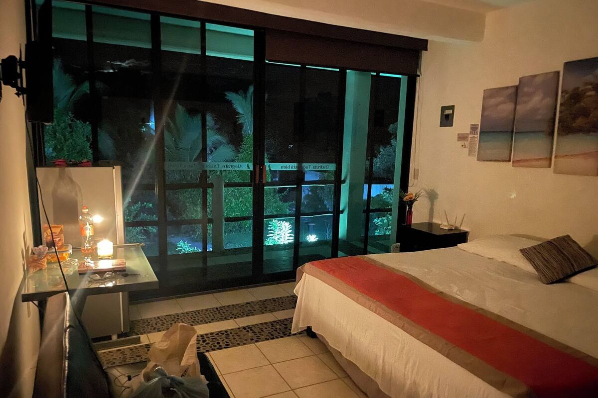 A hotel room with a bed, a desk, a chair, a table with a lamp, a vase with flowers, and a view through large windows.